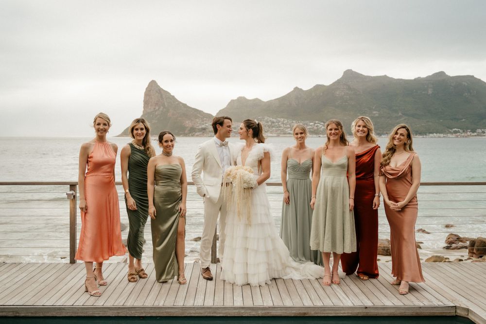 Wedding party group photo on wooden deck overlooking mountain and ocean landscape.