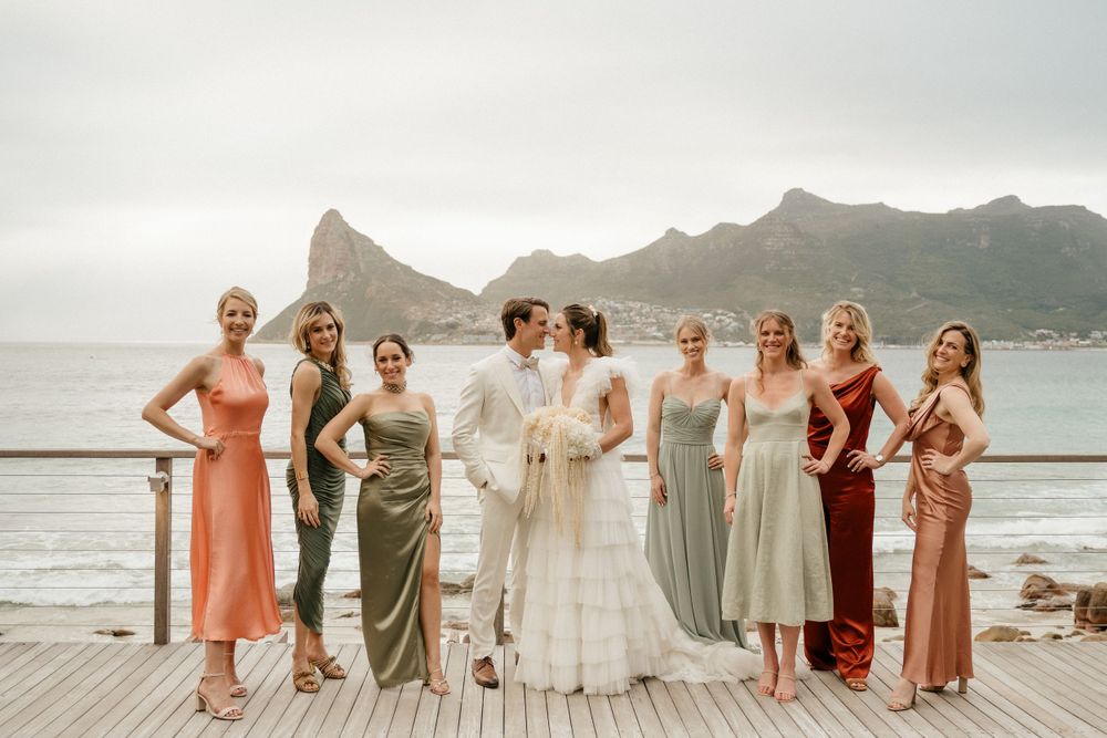 Bridal party in elegant dresses poses on wooden deck with coastal mountain view.