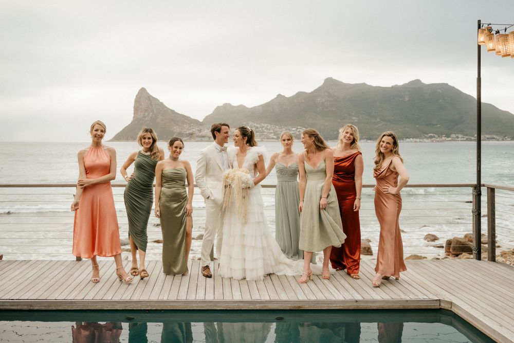 Wedding party poses together on oceanfront deck with mountain backdrop during sunset celebration.