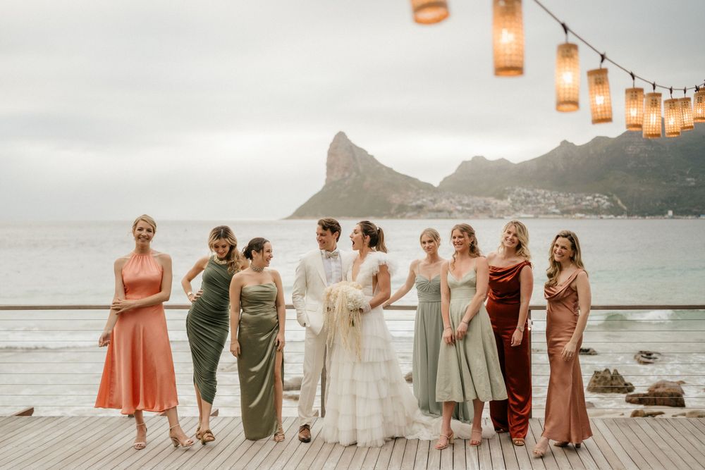 Wedding group photo on oceanfront deck with string lights and mountain backdrop at dusk.