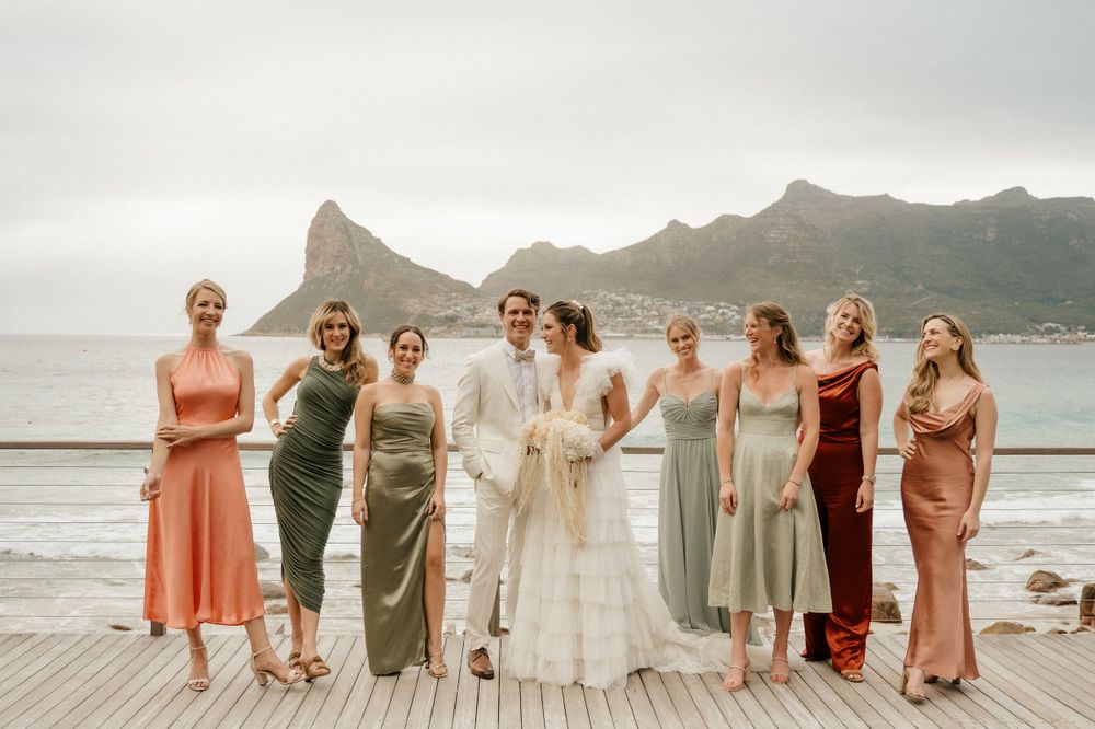 Group photo of wedding party in coordinated dresses on dock with mountain scenery.