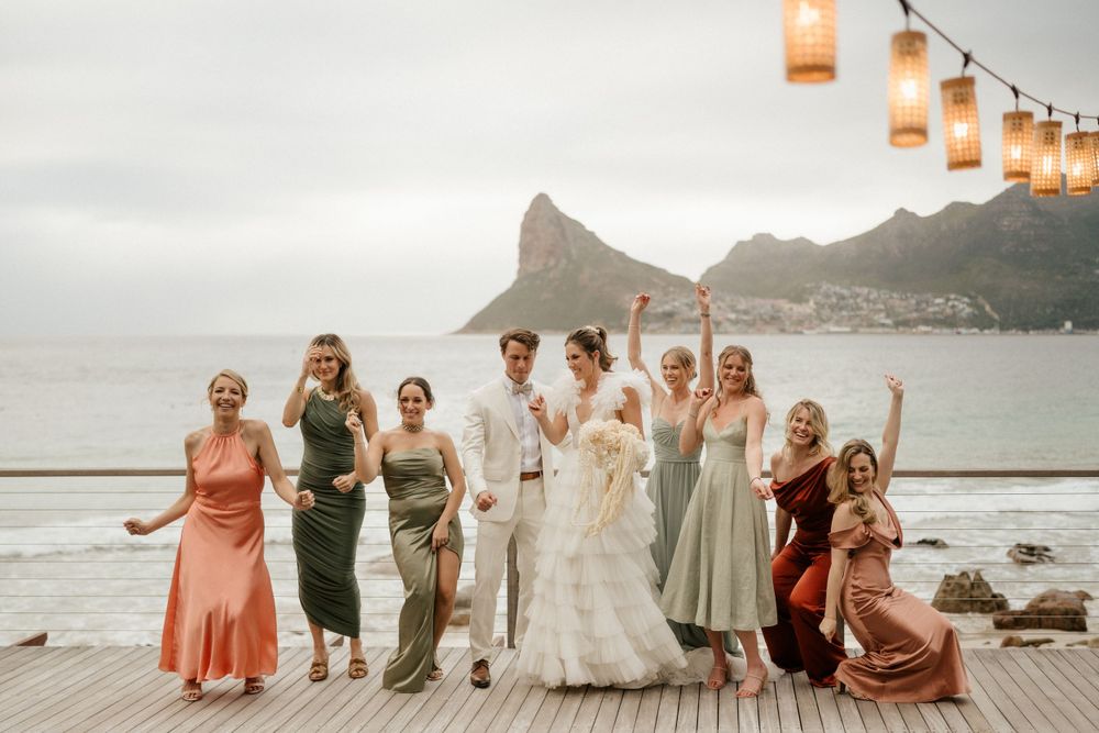 Wedding group poses joyfully on a seaside deck with string lights and mountain backdrop at dusk.