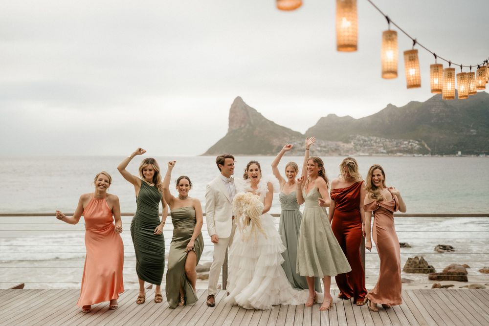 Joyful wedding party celebrates on a wooden deck with mountain scenery and string lights at sunset.