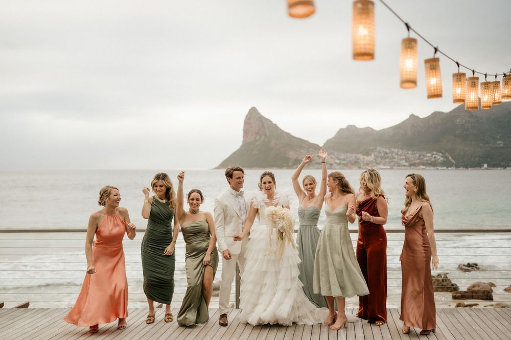 Wedding group poses on wooden deck with string lights and mountain coastline in background.