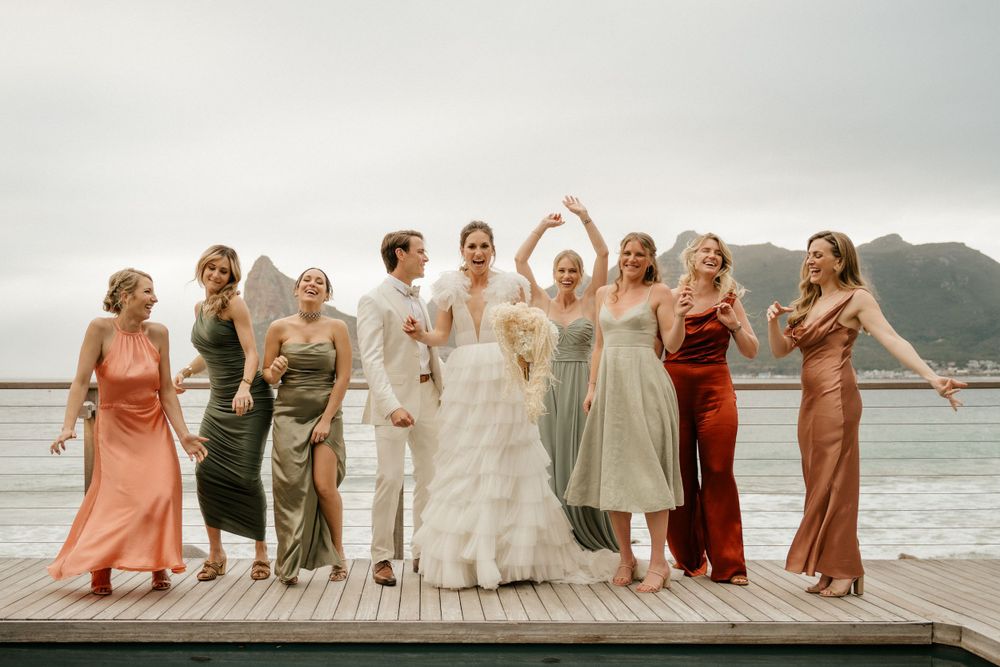 Wedding party stands together on a wooden dock wearing mix of coral, sage, and neutral-toned dresses against mountain backdrop.