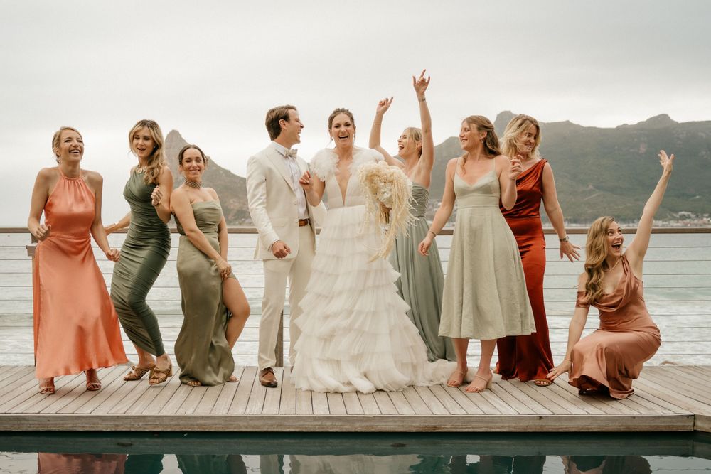 Wedding party poses joyfully on wooden deck with ocean and mountain backdrop.
