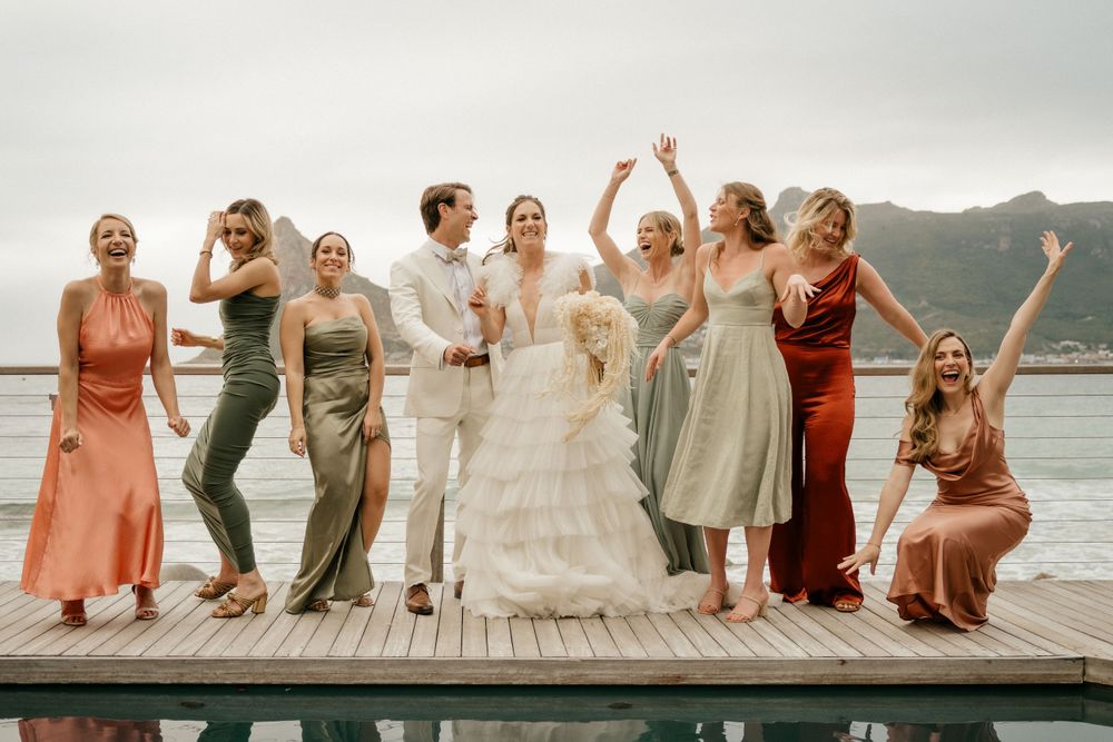 Wedding party stands joyfully on a dock against mountain backdrop in mix of coral and sage dresses.