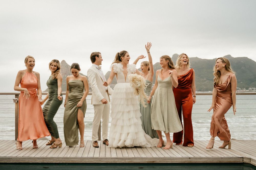 Wedding party laughs together on oceanfront deck with mountain backdrop.