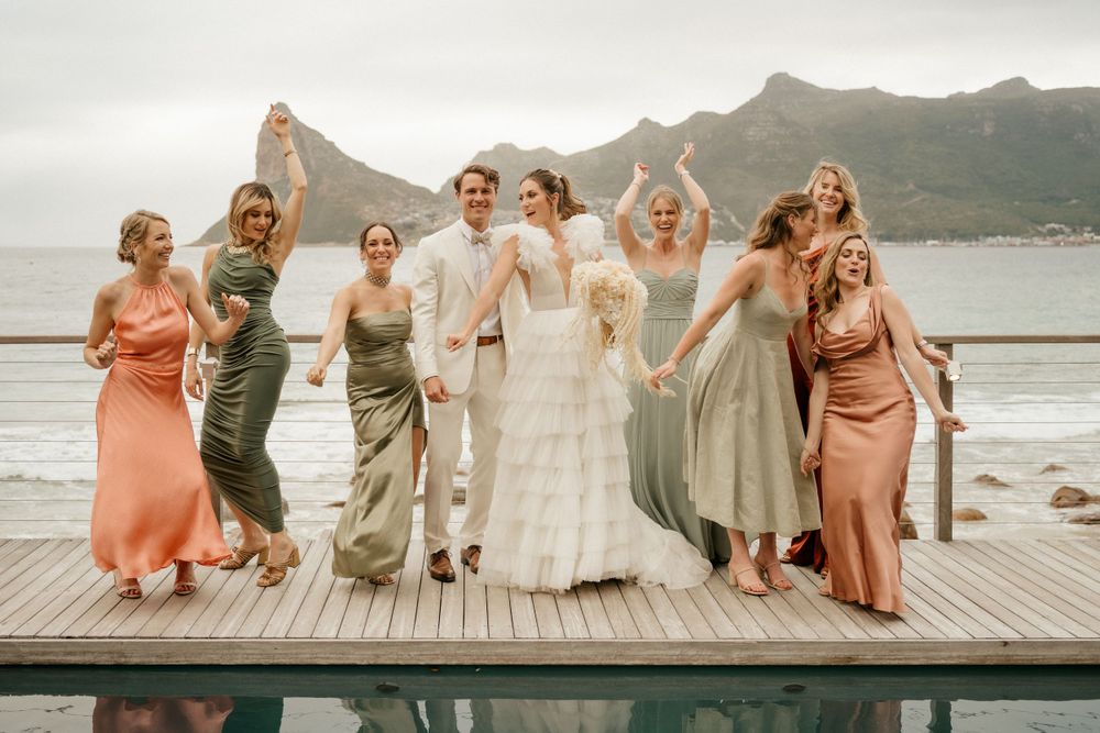 Wedding party poses on a wooden deck with mountain and ocean views wearing coordinated pastel dresses.