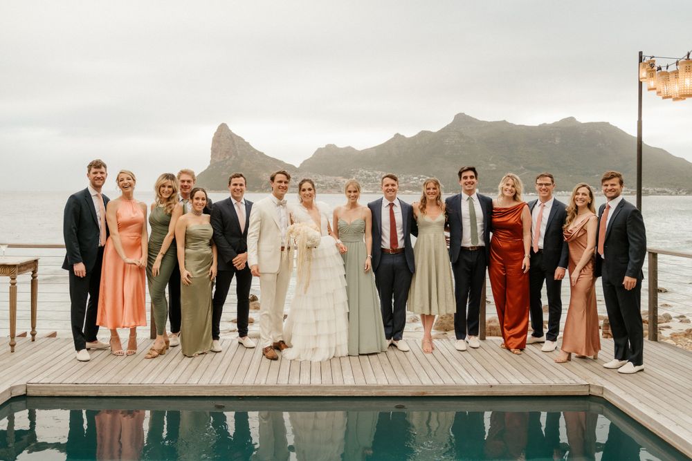 Wedding party lined up on deck beside infinity pool with mountain landscape in background.
