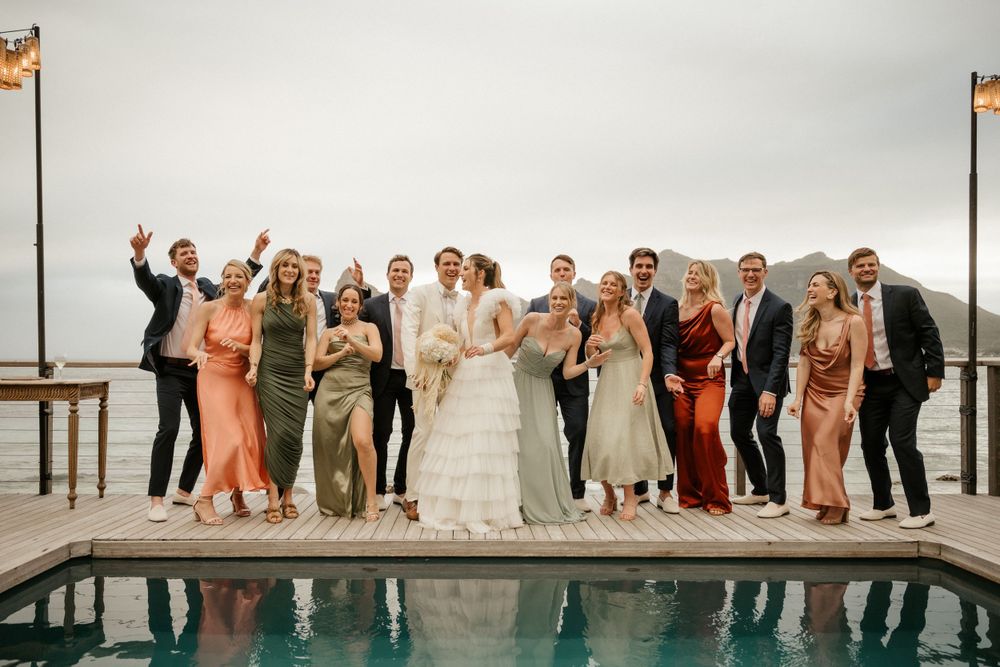 Wedding party celebrates joyfully while standing along the edge of an infinity pool with mountain views.