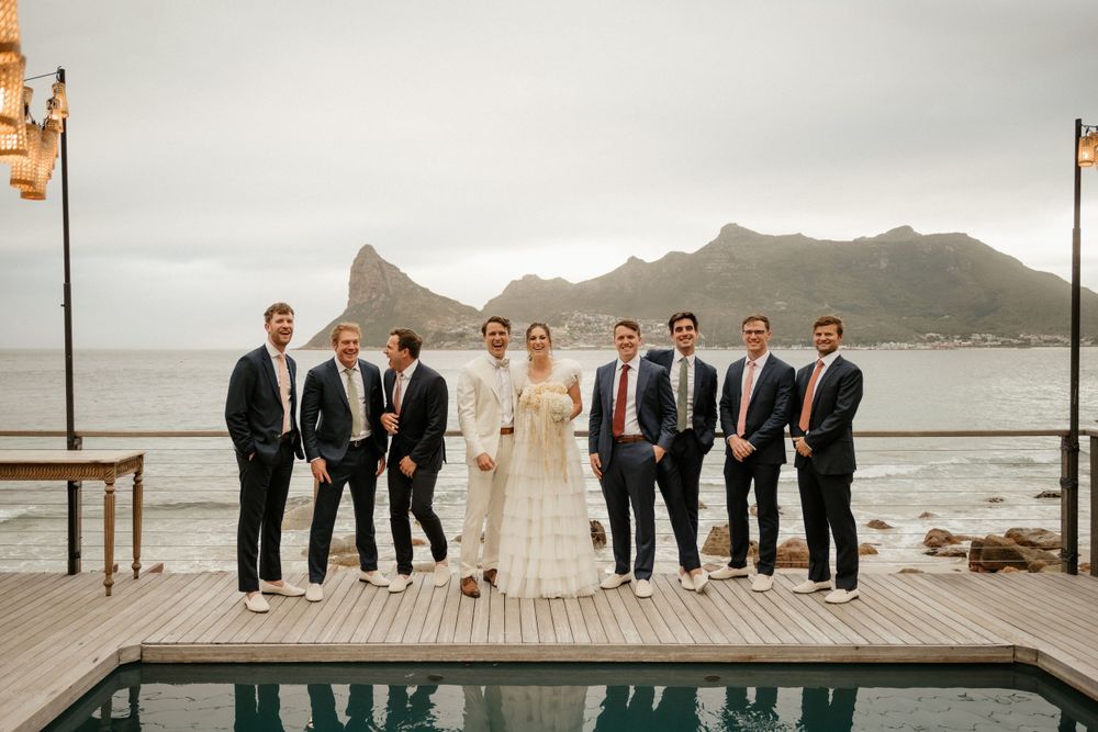 Wedding party stands on seaside deck with dramatic mountain landscape in background.