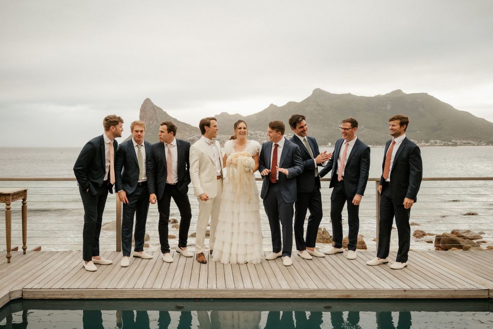 Wedding party posed on wooden deck with mountain and ocean backdrop.