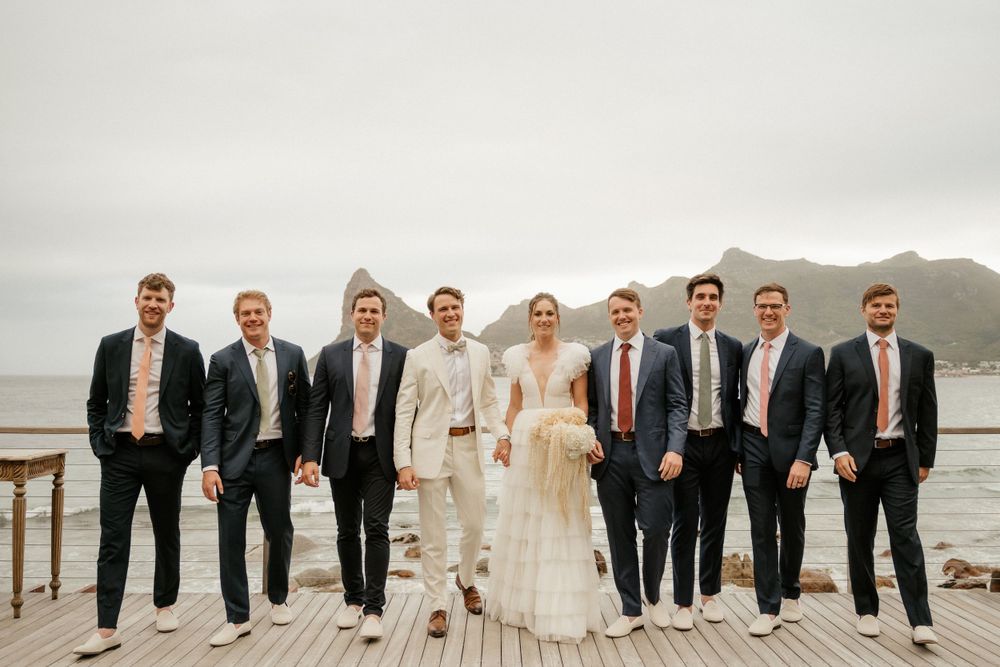 Wedding party in formal attire poses on wooden deck overlooking misty mountain landscape and water.