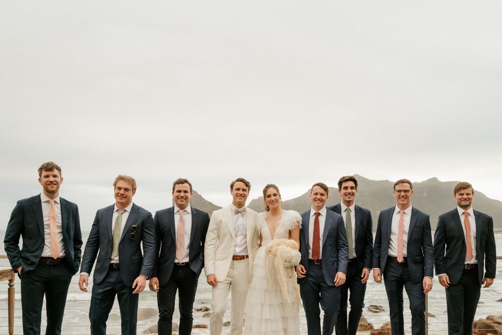 Wedding party stands together on scenic mountain overlook wearing dark suits and white gowns.