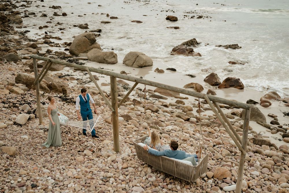 Two people rest on a wooden swing overlooking a rocky beach landscape.