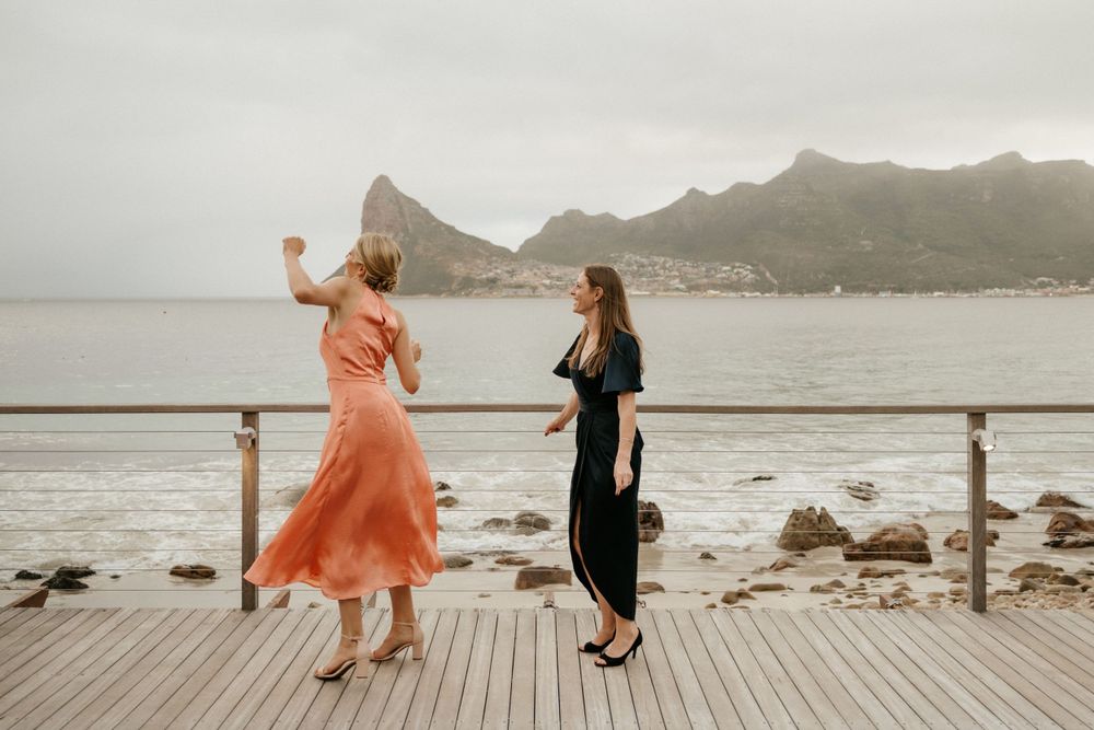 Two wedding guests dancing on oceanfront deck with mountains in background.