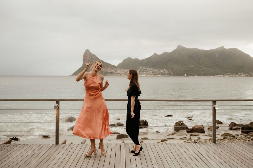 Two people share a moment on a coastal boardwalk with mountains in the background.
