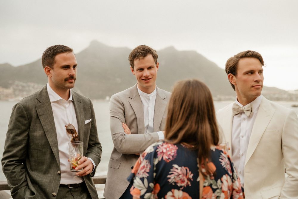 A small gathering of people in light colored suits chat during an outdoor event with mountains in the background.