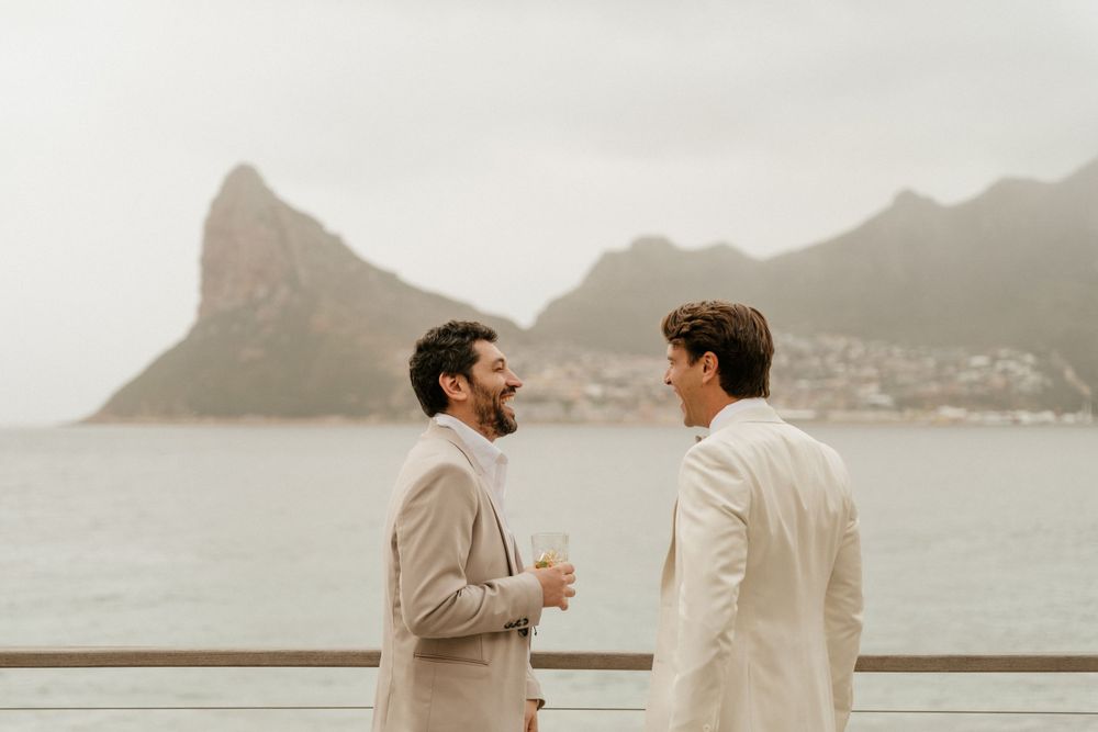 Two people in light colored suits having a conversation on a balcony overlooking coastal mountains.
