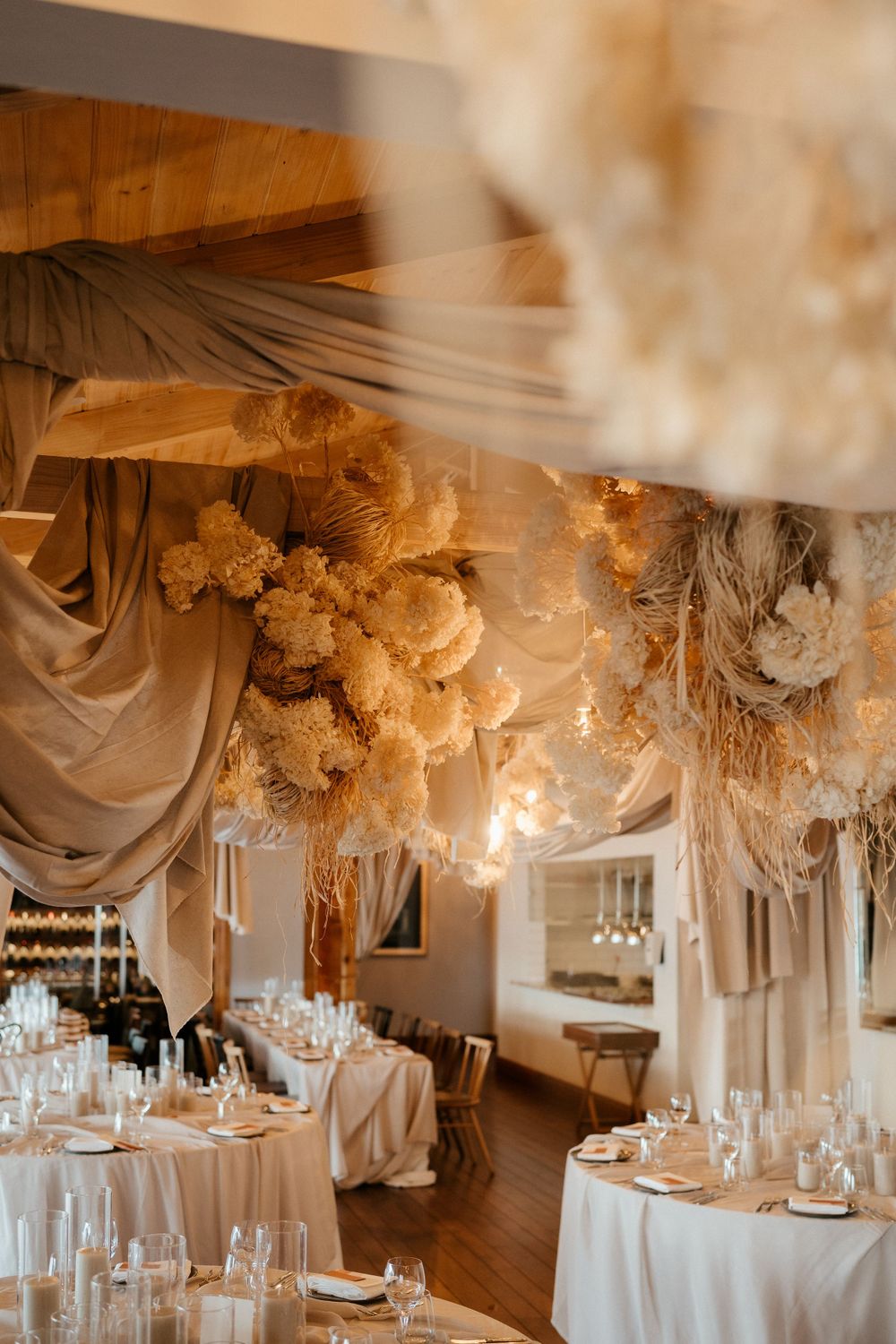 Elegant wedding reception venue with draped fabric ceiling, dried flowers, and white round tables with place settings.