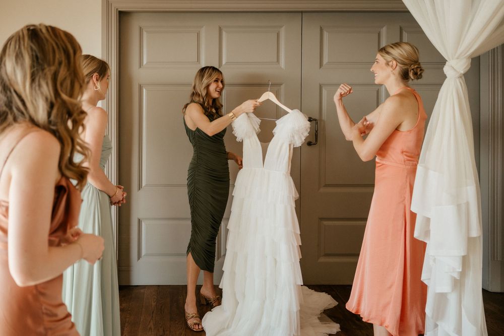 Wedding preparations showing bridesmaids admiring a white wedding gown hanging in a room.