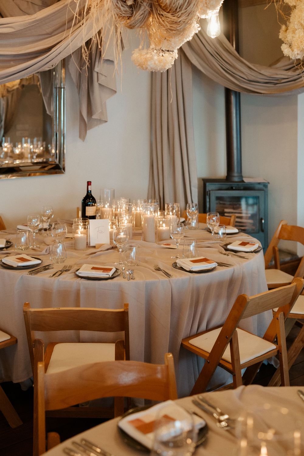 Elegant reception table with draped fabric decor, candles and wooden chairs around a white tablecloth.