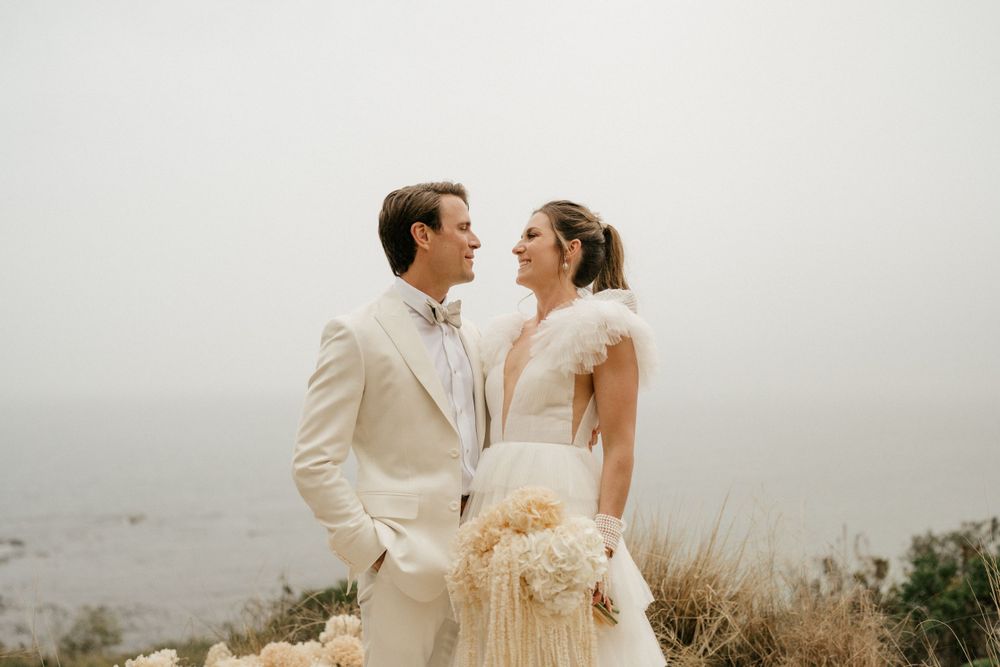 Wedding couple sharing an intimate moment against a misty coastal backdrop in matching cream outfits.