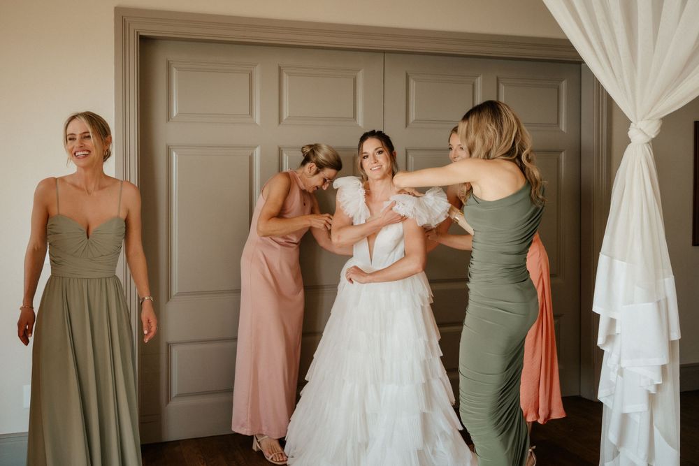 Wedding party helps with final dress preparations in a light-filled room.