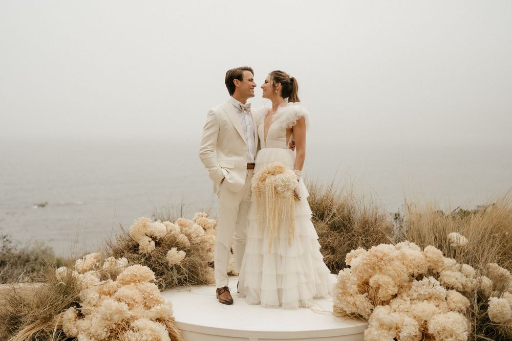 Romantic coastal wedding portrait with pampas grass decor and misty ocean backdrop.
