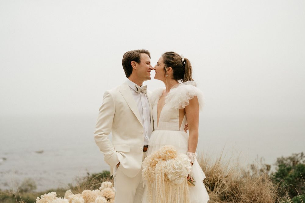 Couple in cream-colored wedding attire share a joyful moment against a foggy landscape.