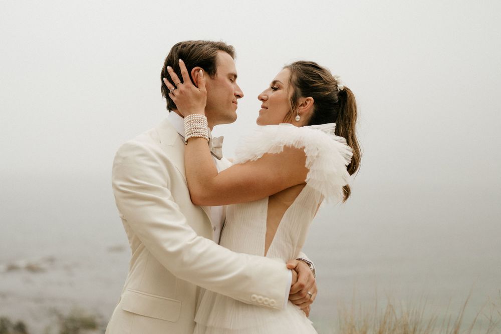 Romantic embrace between couple in cream-colored wedding attire against misty backdrop.