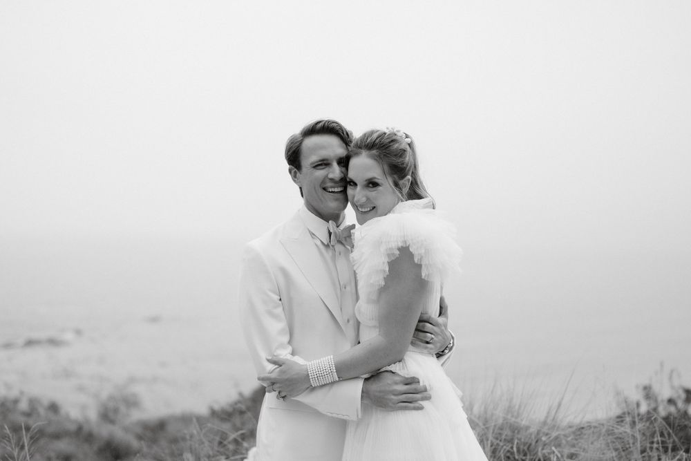 Couple in white attire embrace during foggy coastal wedding photoshoot.