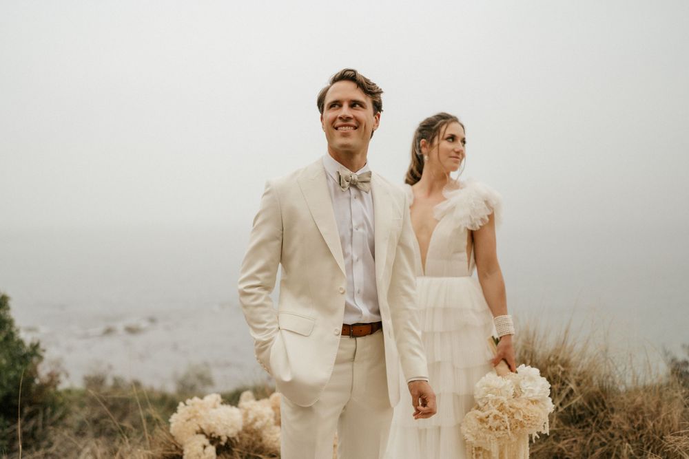 Couple in light colored wedding attire standing against misty coastal backdrop.