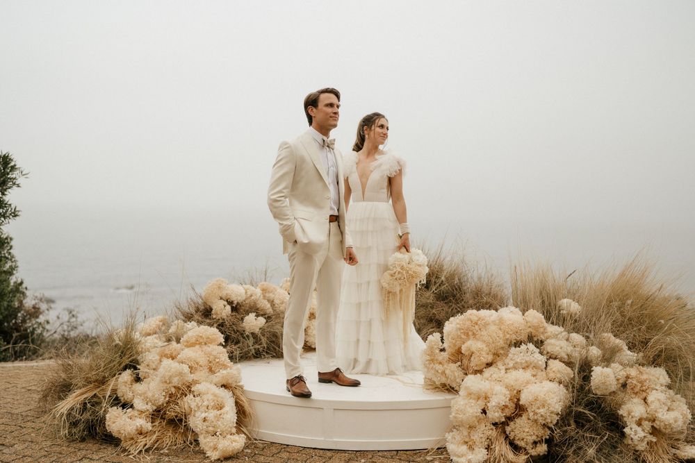 Couple in white wedding attire standing on platform surrounded by cream colored floral arrangements.
