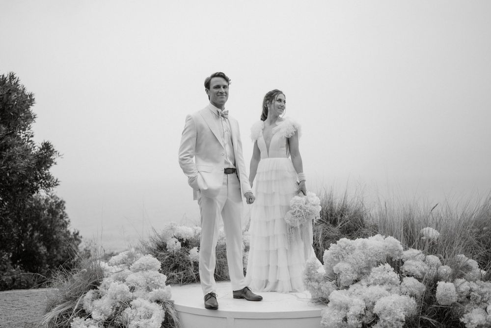 Black and white photo of couple in elegant white attire walking along floral-lined path.