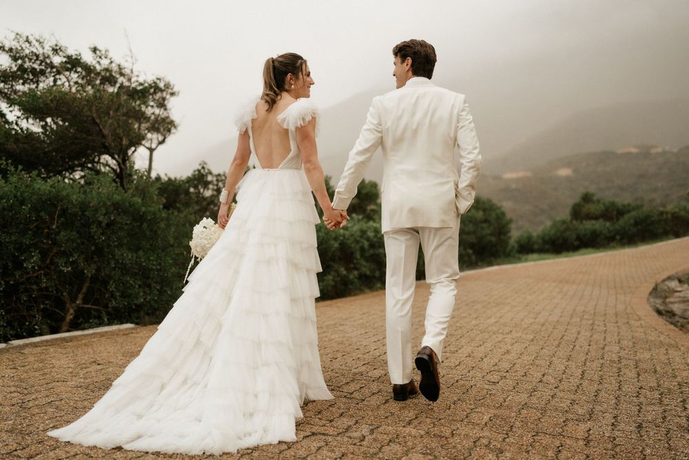 Newlyweds walking hand in hand on a brick path with mountain views.
