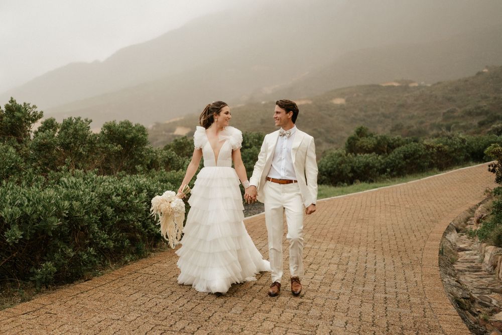 Newlyweds walk hand in hand on stone path with misty mountain views.