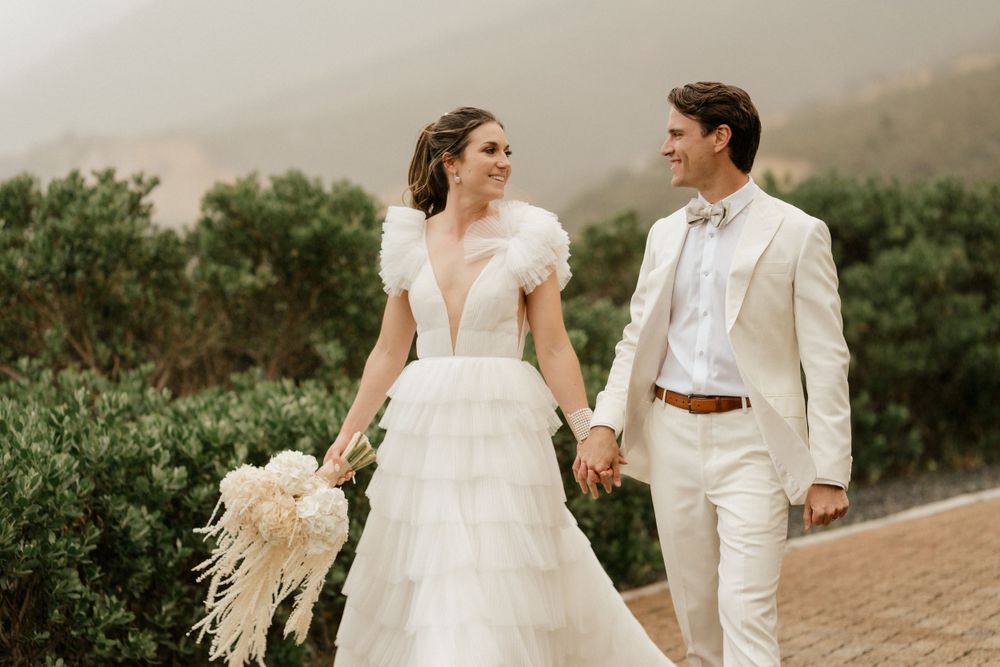 Couple walks hand in hand on coastal path wearing elegant white wedding attire.