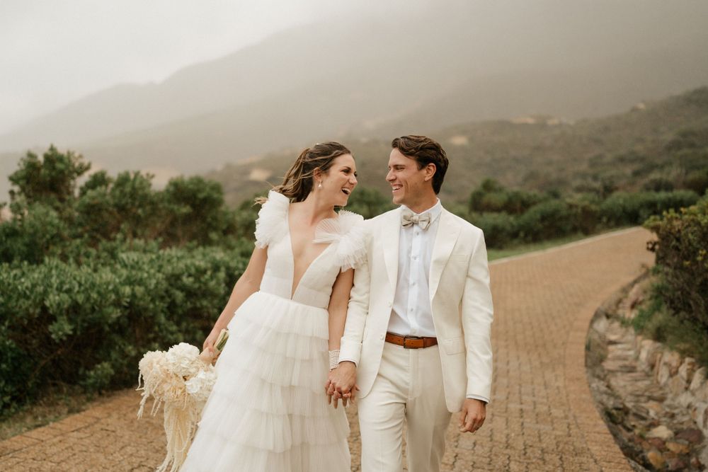 A couple in elegant white wedding attire walks along a winding path with mountain views.