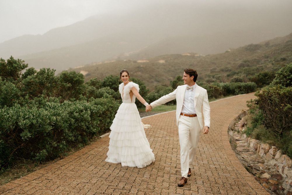 Couple in wedding attire strolls along a scenic mountain pathway.