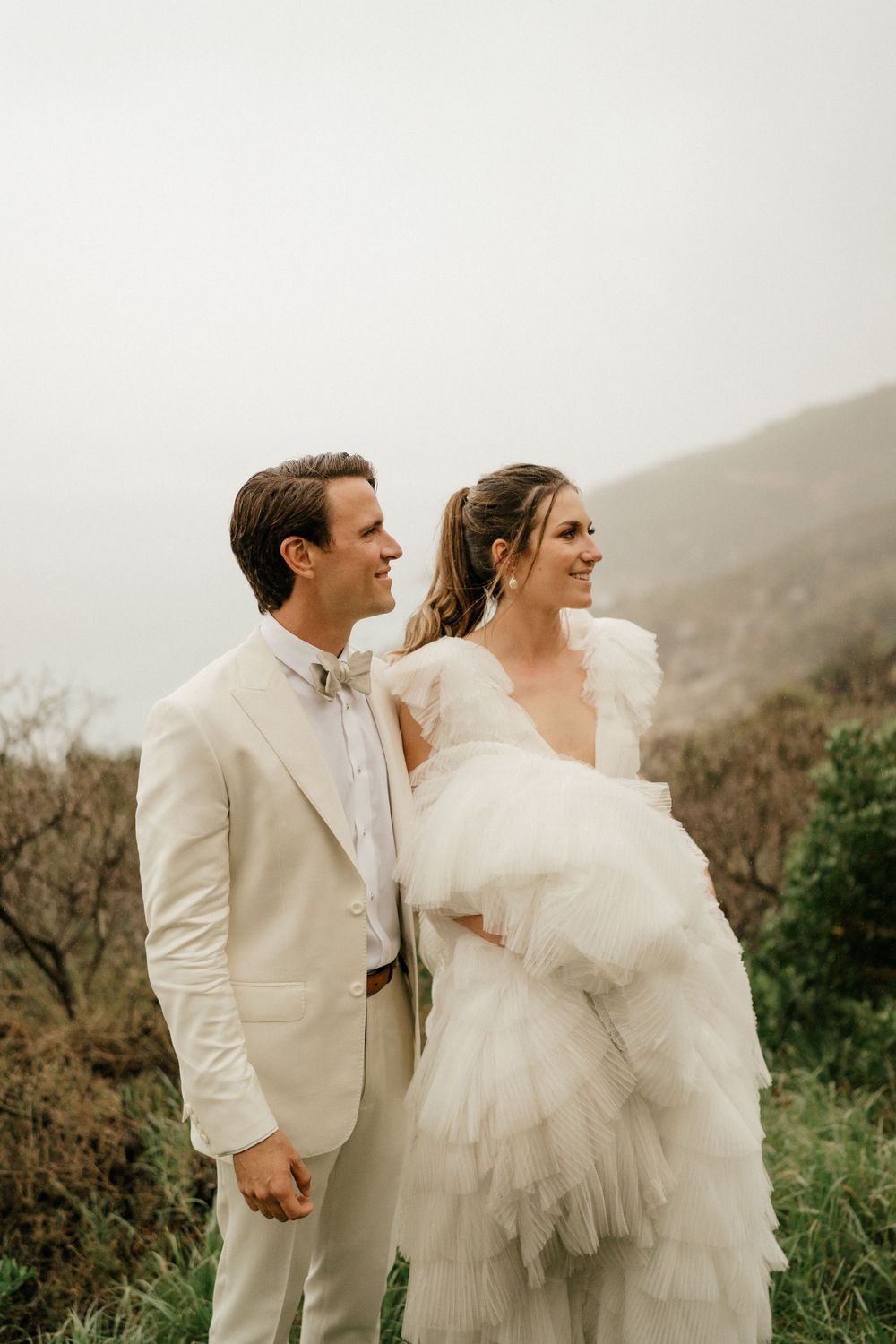 Newlyweds in light-colored attire share a moment on a misty mountainside.
