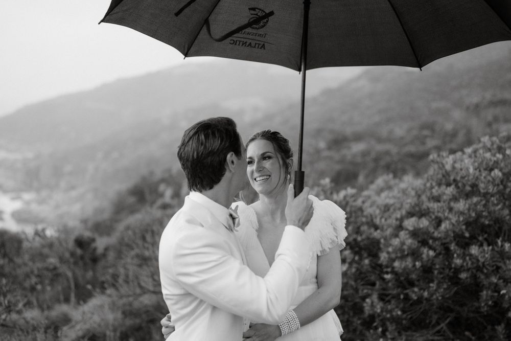 A couple shares a tender moment under a black umbrella against a misty mountain backdrop.