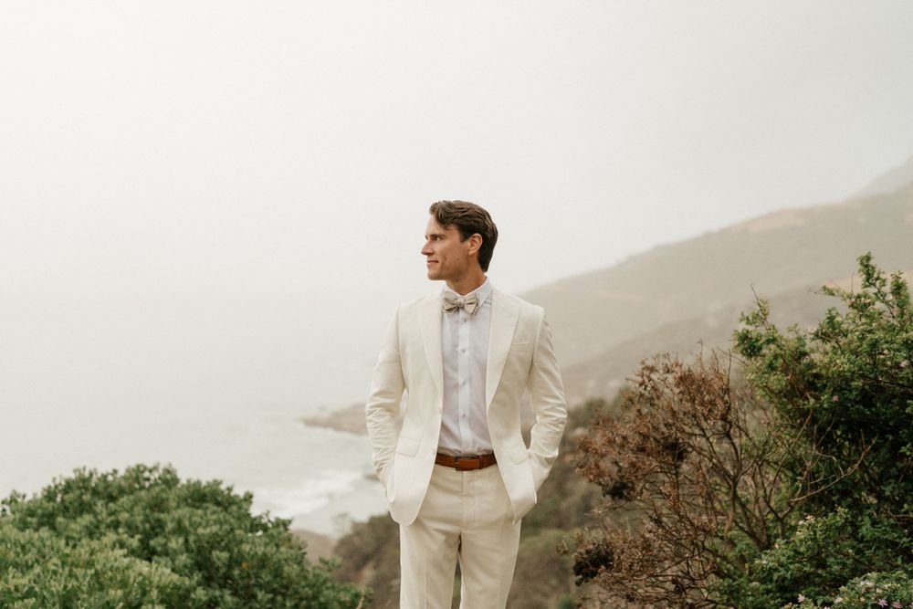 A person in a cream-colored suit stands thoughtfully against a misty mountain backdrop with greenery.