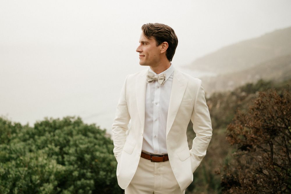 Person in white suit jacket and bow tie poses against misty mountain landscape.