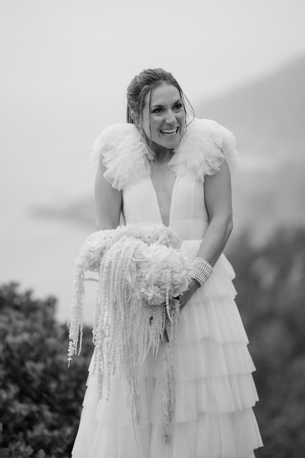 A joyful bride in a tiered white wedding dress with ruffled details poses outdoors against a misty mountain backdrop.