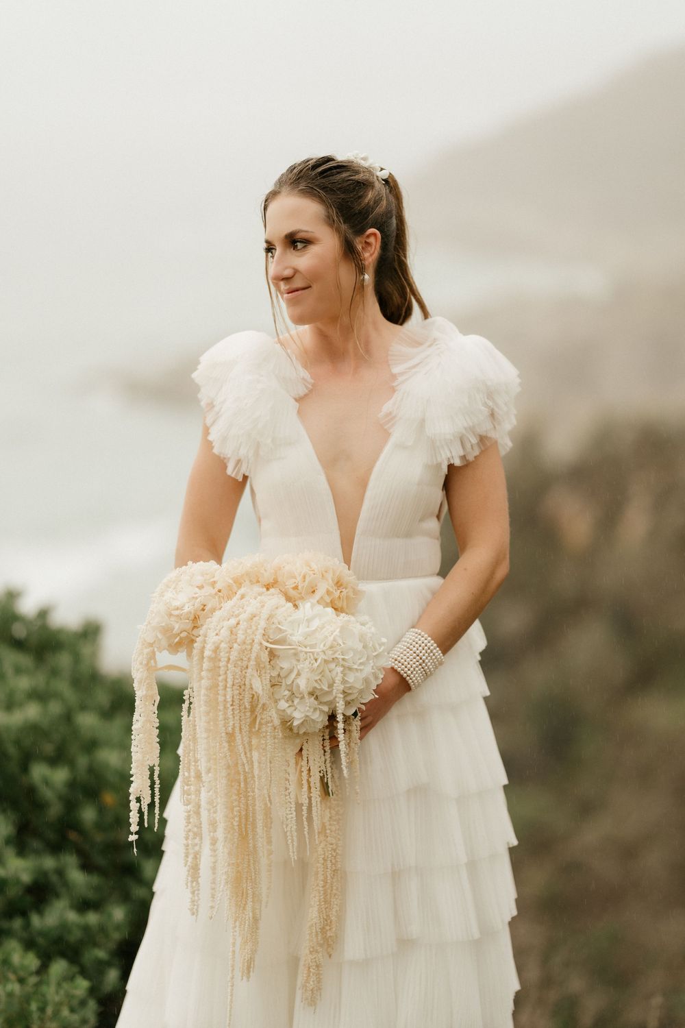 A bride in a layered white wedding dress with ruffled cap sleeves holds a cream-colored bouquet against a misty mountain backdrop.