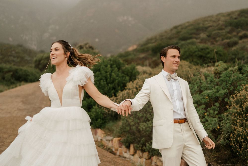 Newlyweds in white attire hold hands while walking on mountain trail during sunset photoshoot.