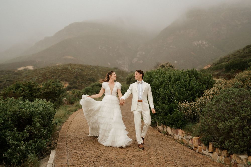 A couple walks hand in hand on a misty mountain path in wedding attire.