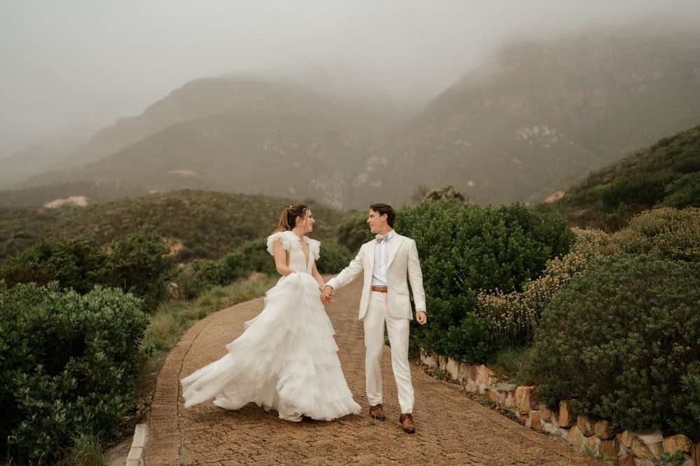 Newlyweds walk hand in hand on a misty mountain path with flowing wedding dress.
